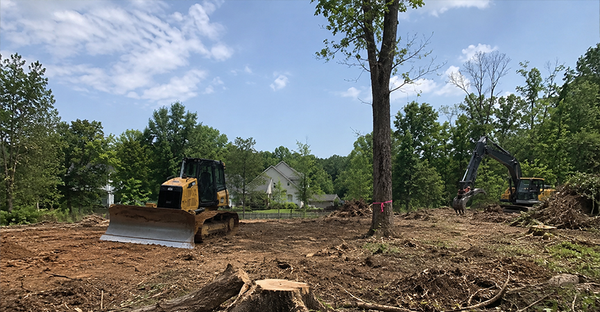 Heavy construction equipment, including a bulldozer and an excavator, clearing trees and grading a dirt lot for a new home construction site.
